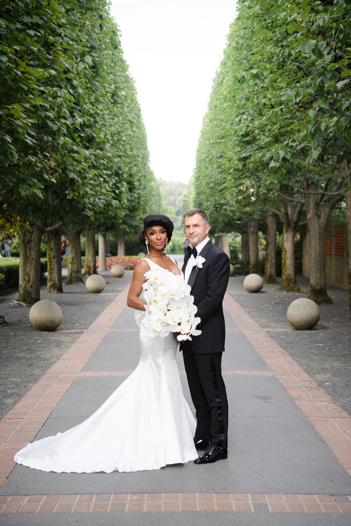 Bride holding bouquet of white orchids posing with groom by tall hedges at the Chicago Botanic Garden